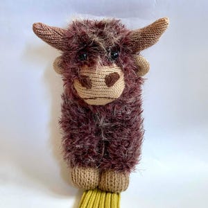 May include: A handmade plush Highland cow toy. The toy features a shaggy, dark brown body, tan horns, and a tan face with black eyes. The cow has tan hooves and a yellow knitted bottom.