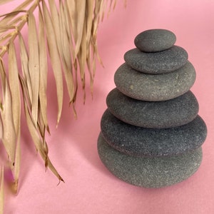 Può includere: Un cumulo di sette pietre grigie bilanciate l'una sull'altra su uno sfondo rosa. Una foglia di palma secca è sullo sfondo.