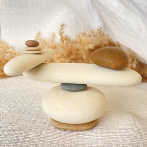Puede incluir: Una escultura equilibrada compuesta por piedras lisas y redondeadas apiladas en una disposición precaria. La base consiste en una piedra grande de color beige claro sobre una piedra ocre, rematada por un guijarro gris oscuro y una piedra larga.