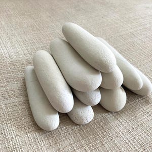 Puede incluir: Una colección de piedras oblongas lisas de color gris claro. Las piedras están apiladas y dispuestas, mostrando sus formas redondeadas y superficies texturizadas. La paleta de colores neutros y las formas naturales sugieren un tema de spa o decoración del hogar.