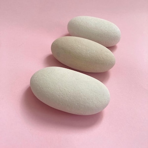Puede incluir: Tres piedras lisas de forma ovalada en tonos blancos y beige sobre un fondo rosa. Las piedras tienen una superficie texturizada y están dispuestas en diagonal. Probablemente decorativas.