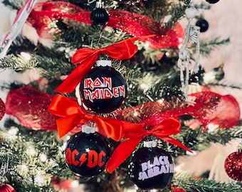 May include: A decorated Christmas tree with black ornaments featuring the logos of rock bands like Metallica, Iron Maiden, AC/DC, and Black Sabbath. The ornaments are accented with red bows and glittery ribbon.