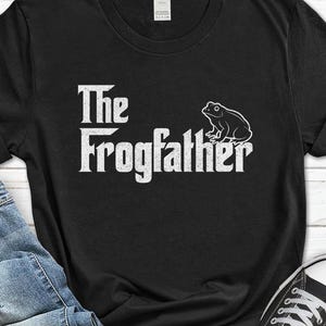 May include: Black t-shirt with the text "The Frogfather" in a white, stylized font, accompanied by a white outline drawing of a frog. The shirt is made of a soft material.