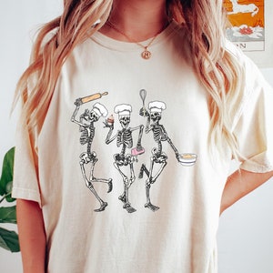 Op de afbeelding: Een wit T-shirt met een afbeelding van drie skeletten die verkleed zijn als koks en aan het bakken zijn. De skeletten houden een deegroller, een cupcake, een mengkom en een garde vast.