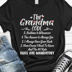 May include: Black t-shirt with the text "The Grandma Code" in white and floral lettering. The shirt lists rules such as "Hugs Are Mandatory" and "Bedtime Is Whenever". A casual, comfortable tee.