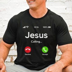 May include: A black t-shirt with a white graphic of a phone screen. The screen displays "Jesus Calling..." with a red "Decline" button and a green "Accept" button. The phone screen also shows a signal strength indicator, the time 03:16, and a battery indicator at 100%.