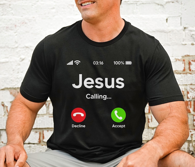 May include: A black t-shirt with a white graphic of a phone screen. The screen displays "Jesus Calling..." with a red "Decline" button and a green "Accept" button. The phone screen also shows a signal strength indicator, the time 03:16, and a battery indicator at 100%.
