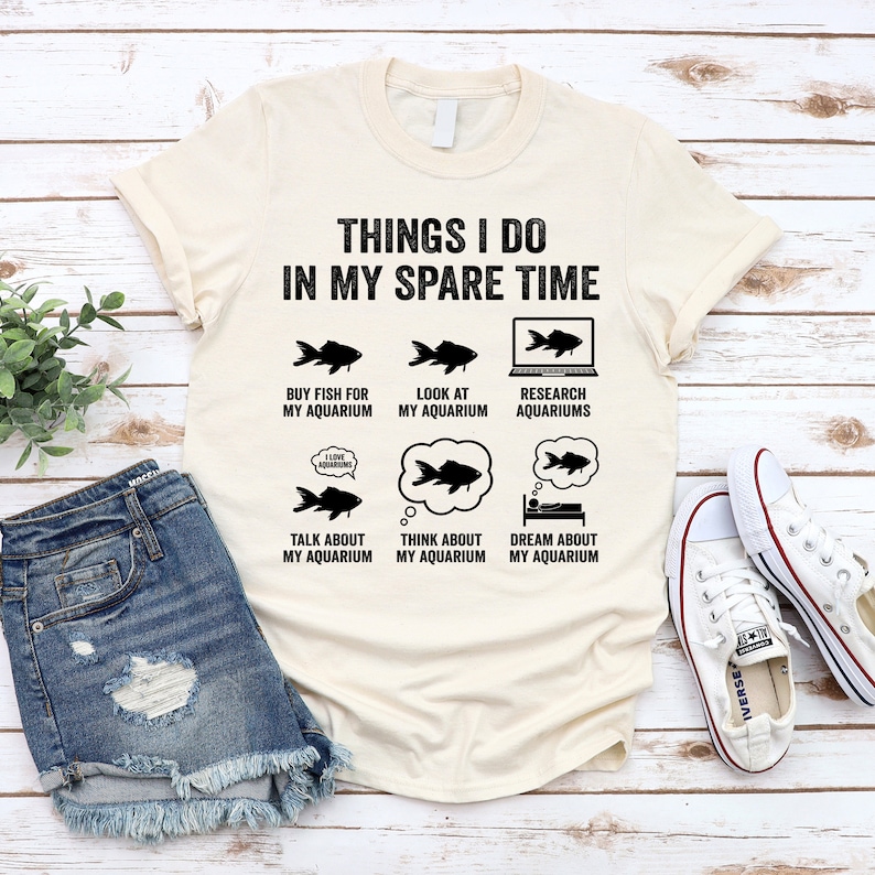 May include: A beige t-shirt with black text that reads "Things I do in my spare time". The text is surrounded by six black and white illustrations of fish and a computer screen. The illustrations depict activities related to aquariums, such as buying fish, looking at aquariums, researching aquariums, talking about aquariums, thinking about aquariums, and dreaming about aquariums.