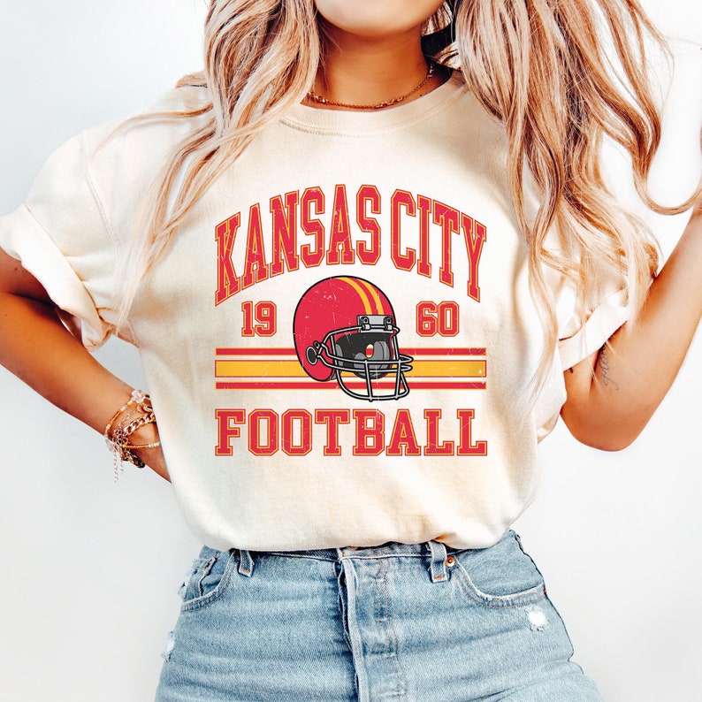 K&ouml;nnte beinhalten: Ein wei&szlig;es T-Shirt mit einer Grafik im Vintage-Stil eines Footballhelms und dem Text "Kansas City Football" mit den Zahlen "19" und "60" auf beiden Seiten des Helms.