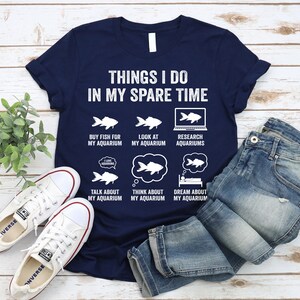 May include: A navy blue t-shirt with a white graphic that says "Things I do in my spare time" and lists activities related to aquariums. The graphic includes illustrations of fish, a computer screen, and a sleeping person.