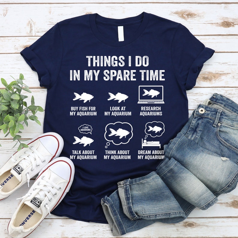 May include: A navy blue t-shirt with a white graphic that says "Things I do in my spare time" and lists activities related to aquariums. The graphic includes illustrations of fish, a computer screen, and a sleeping person.