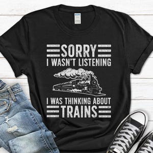 May include: Black t-shirt with white text and a train graphic. The text reads "SORRY I WASN'T LISTENING I WAS THINKING ABOUT TRAINS". The graphic is a detailed illustration of a steam train.