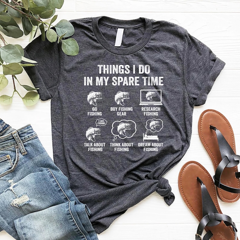 May include: A dark heather grey t-shirt with a white graphic design that says "Things I do in my spare time". The graphic features a series of illustrations of fish and text related to fishing.