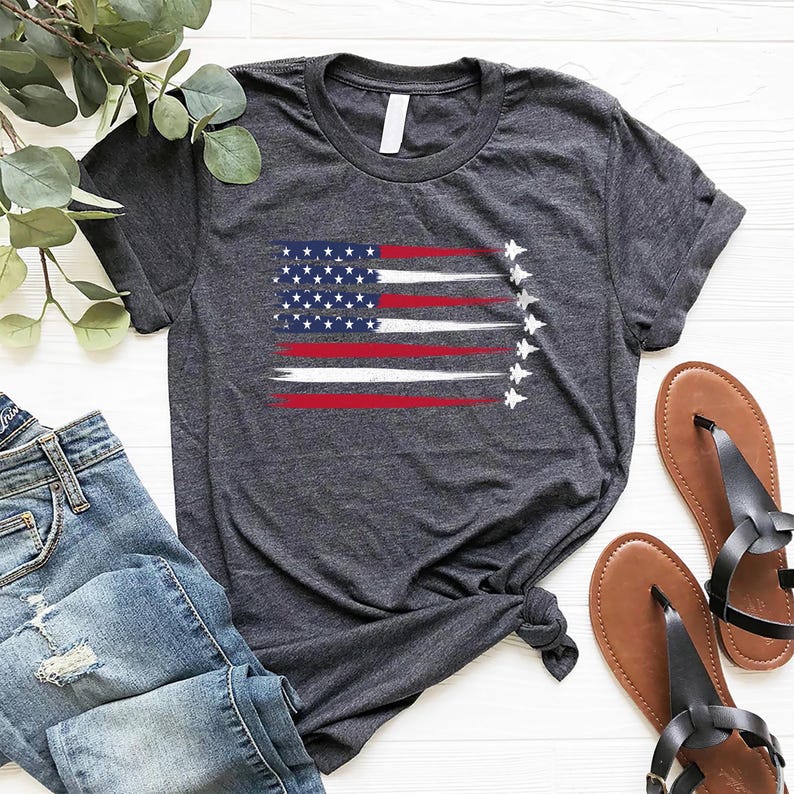 Puede incluir: Una camiseta gris oscuro jaspeada con un dise&ntilde;o de bandera estadounidense desgastada y siluetas blancas de aviones de combate volando en formaci&oacute;n.