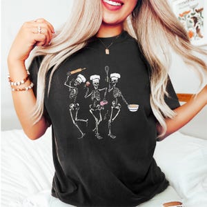 Op de afbeelding: Zwart T-shirt met een afbeelding van drie skeletten die als koks zijn verkleed. De skeletten houden een deegroller, een garde en een kom vast. De tekst "Bake 'em Dead" staat op het shirt gedrukt.