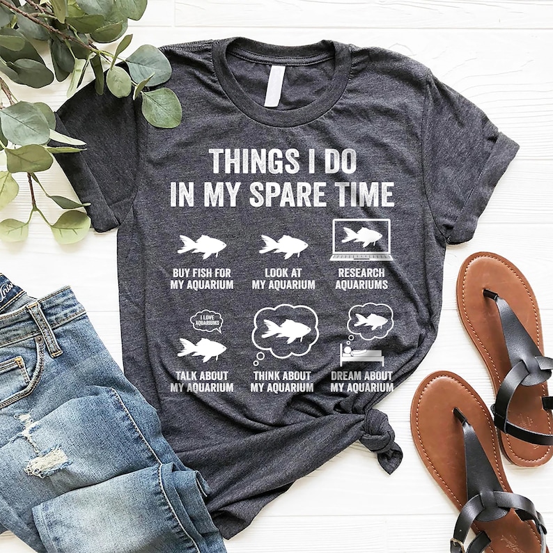 May include: A dark gray t-shirt with the text "Things I do in my spare time" and illustrations of fish, aquariums, and a person sleeping. The text "I love aquariums" is in a speech bubble.