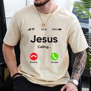 May include: A beige t-shirt with a black graphic of a phone screen. The screen displays the text "Jesus Calling..." with two buttons: "Decline" and "Accept".