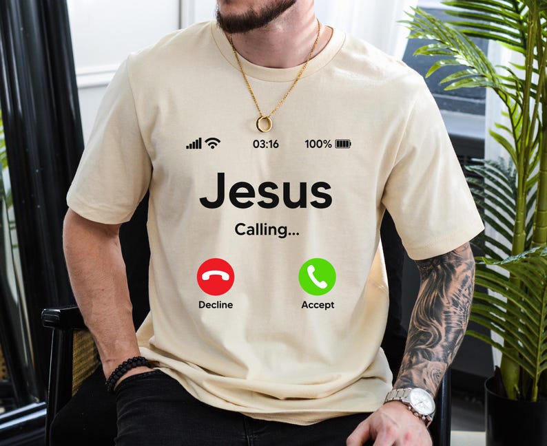 May include: A beige t-shirt with a black graphic of a phone screen. The screen displays the text "Jesus Calling..." with two buttons: "Decline" and "Accept".