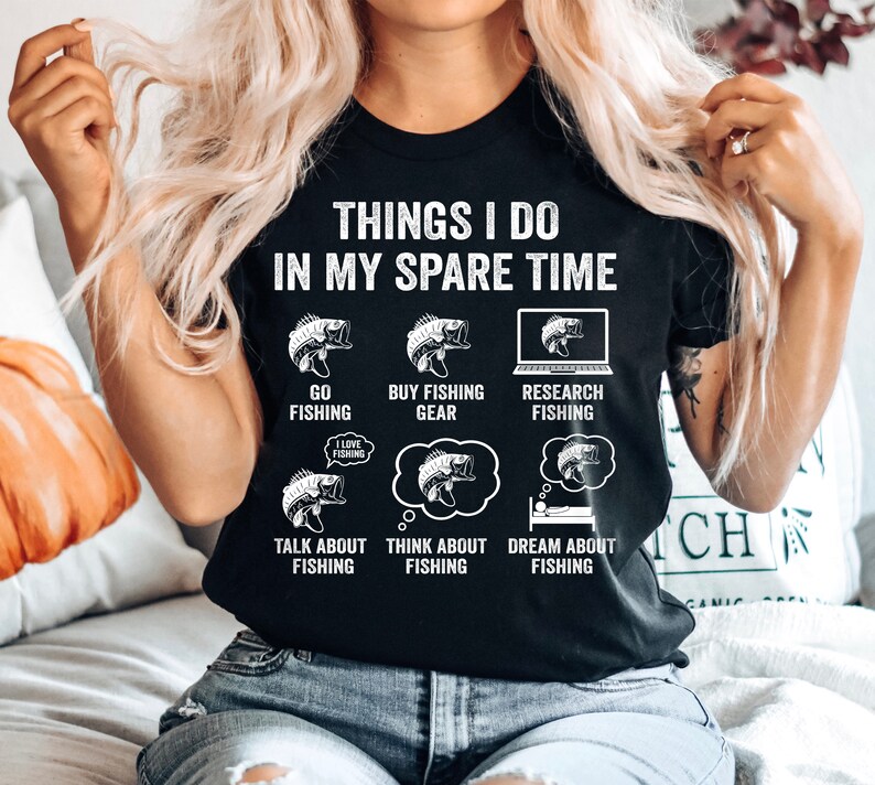 May include: Black t-shirt with white text that reads "Things I do in my spare time". The text is surrounded by illustrations of fish and fishing-related activities, including "Go Fishing", "Buy Fishing Gear", "Research Fishing", "Talk About Fishing", "Think About Fishing", and "Dream About Fishing".