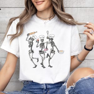 Op de afbeelding: Wit T-shirt met een afbeelding van drie skeletten die als koks zijn verkleed. De skeletten houden een deegroller, een garde, een cupcake en een kom vast.