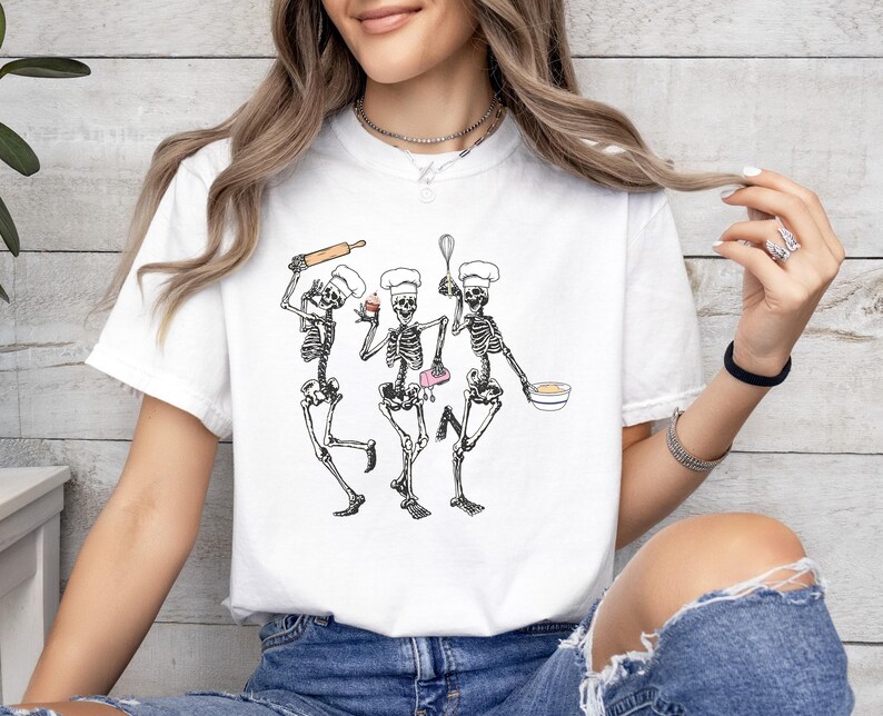 Op de afbeelding: Wit T-shirt met een afbeelding van drie skeletten die als koks zijn verkleed. De skeletten houden een deegroller, een garde, een cupcake en een kom vast.
