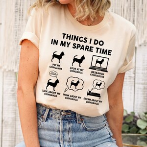 May include: A beige t-shirt with a black graphic that says "Things I do in my spare time" and features a series of illustrations of a chihuahua dog. The illustrations show the dog being petted, being looked at, being researched online, being talked about, being thought about, and being dreamed about.