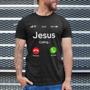 May include: A dark grey t-shirt with a white graphic of a phone screen. The screen displays "Jesus Calling..." with a red decline button and a green accept button.