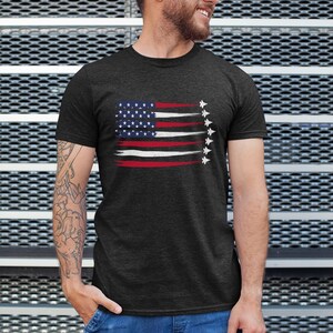 Puede incluir: Una camiseta gris oscuro con un dise&ntilde;o de bandera estadounidense desgastado que presenta una fila de aviones de combate volando en formaci&oacute;n.