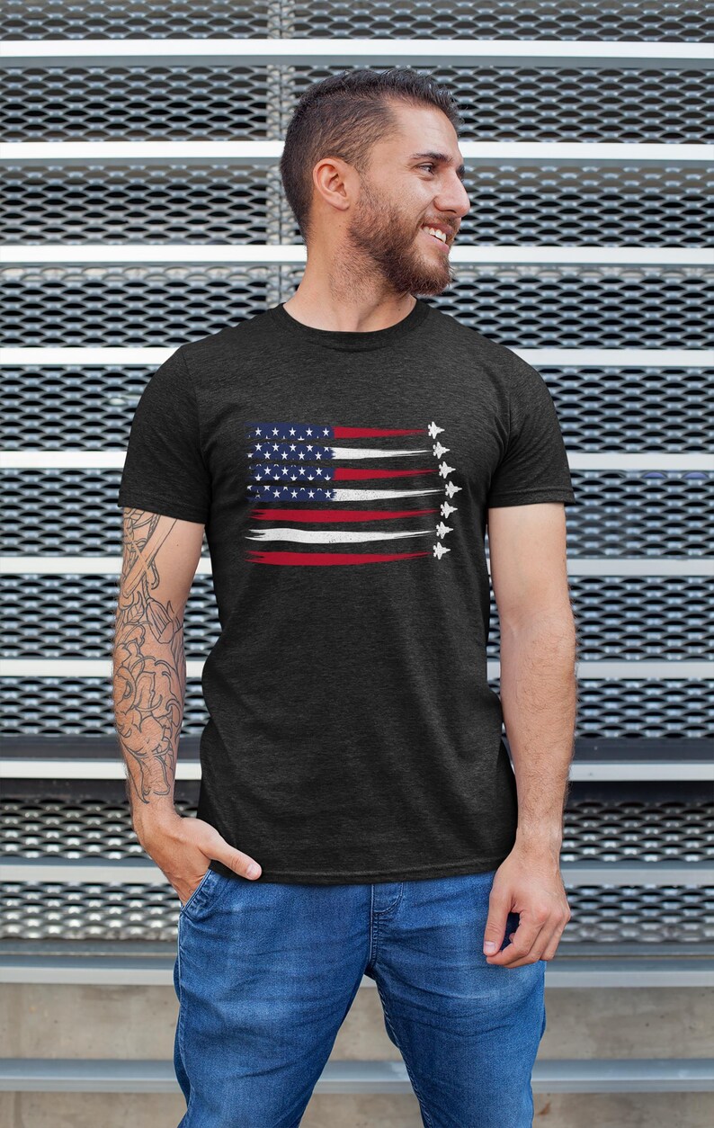 Puede incluir: Una camiseta gris oscuro con un dise&ntilde;o de bandera estadounidense desgastado que presenta una fila de aviones de combate volando en formaci&oacute;n.