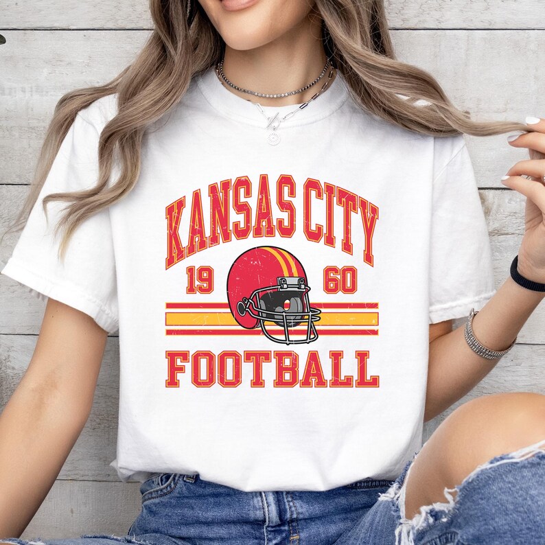 K&ouml;nnte beinhalten: Wei&szlig;es T-Shirt mit einem roten, gelben und schwarzen Grafikdesign. Die Grafik zeigt einen Footballhelm mit dem Text "Kansas City 19 60 Football".