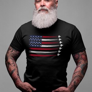 Puede incluir: Un hombre con una camiseta negra con un dise&ntilde;o de bandera estadounidense desgastado y una fila de aviones de combate volando en formaci&oacute;n. Tiene una barba blanca larga y tatuajes en los brazos.