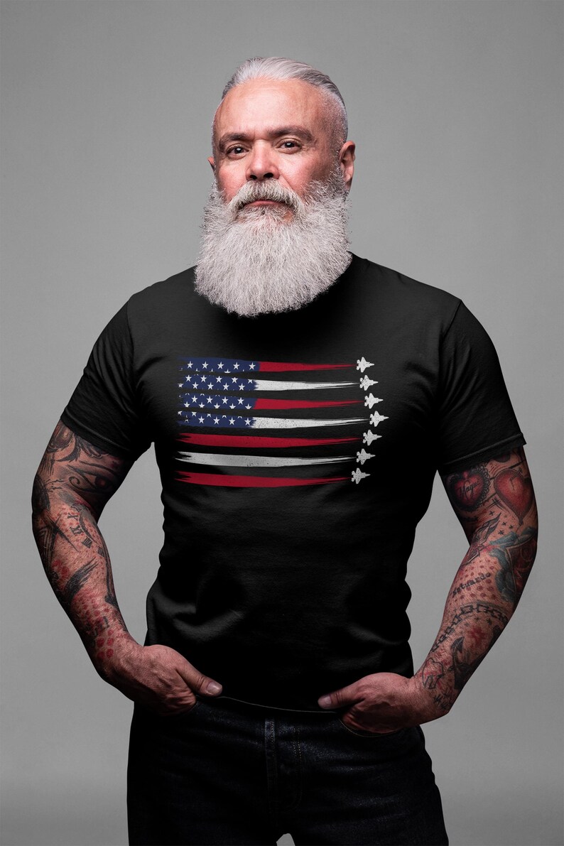 Puede incluir: Un hombre con una camiseta negra con un dise&ntilde;o de bandera estadounidense desgastado y una fila de aviones de combate volando en formaci&oacute;n. Tiene una barba blanca larga y tatuajes en los brazos.
