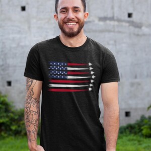 Puede incluir: Una camiseta negra con un dise&ntilde;o de bandera estadounidense desgastada y una fila de aviones de combate volando en formaci&oacute;n. La bandera es roja, blanca y azul.