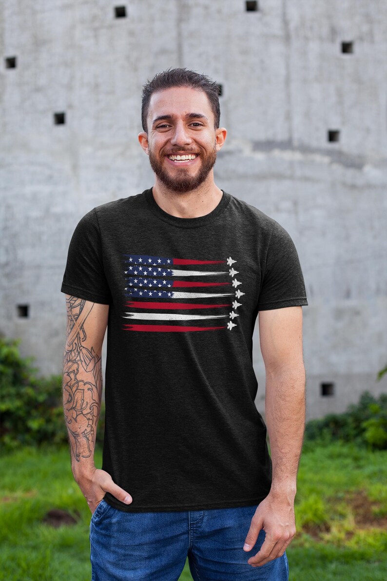 Puede incluir: Una camiseta negra con un dise&ntilde;o de bandera estadounidense desgastada y una fila de aviones de combate volando en formaci&oacute;n. La bandera es roja, blanca y azul.