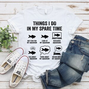 May include: White t-shirt with black text that reads "Things I do in my spare time". The text is surrounded by illustrations of fish and aquariums. The illustrations include the text "Buy fish for my aquarium", "Look at my aquarium", "Research aquariums", "Talk about aquarium", "Think about my aquarium", and "Dream about my aquarium".