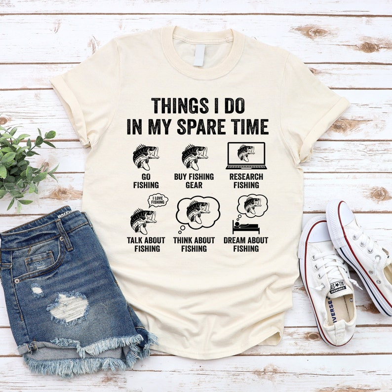 May include: A beige t-shirt with the text "Things I do in my spare time" and a series of illustrations of fish and fishing related activities. The text includes "Go Fishing", "Buy Fishing Gear", "Research Fishing", "Talk About Fishing", "Think About Fishing", and "Dream About Fishing".