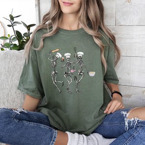 Op de afbeelding: Een groen T-shirt met een afbeelding van drie skeletten die als koks zijn verkleed. De skeletten houden bakgerei en een cupcake vast.