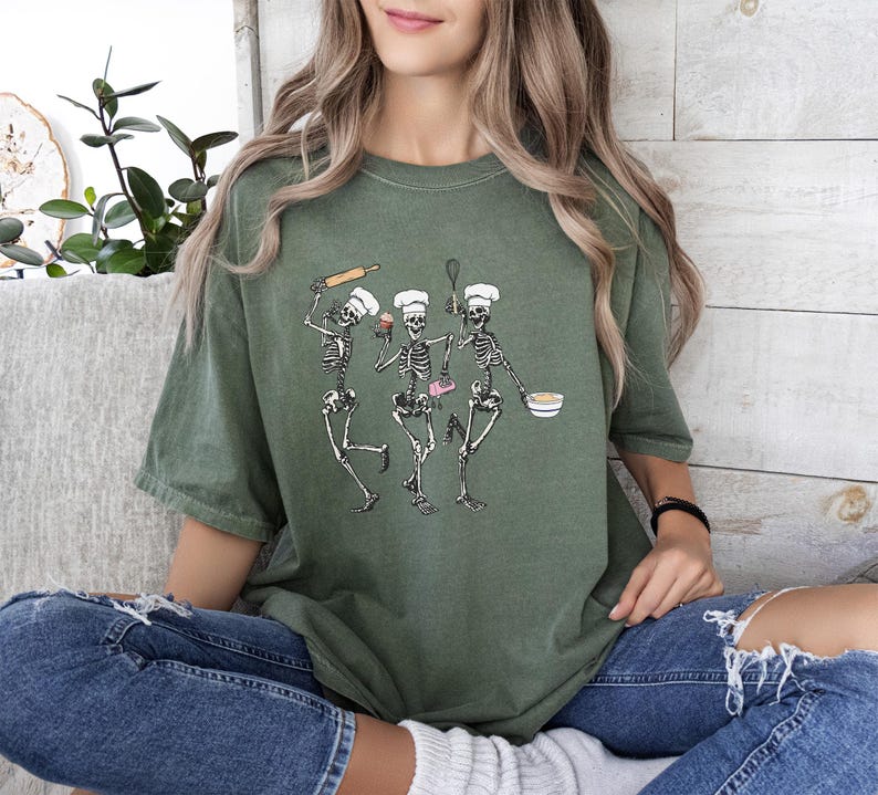 Op de afbeelding: Een groen T-shirt met een afbeelding van drie skeletten die als koks zijn verkleed. De skeletten houden bakgerei en een cupcake vast.