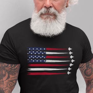 Puede incluir: Una camiseta negra con un dise&ntilde;o de bandera estadounidense desgastado que presenta una fila de aviones de combate volando en formaci&oacute;n.