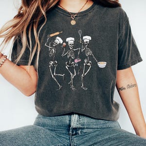 Op de afbeelding: Een donkergrijs T-shirt met een afbeelding van drie skeletten die als koks zijn verkleed. De skeletten houden bakgerei en een cupcake vast. Het shirt heeft een verweerde look.