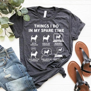 May include: A dark gray t-shirt with the text "THINGS I DO IN MY SPARE TIME" and a series of illustrations of a chihuahua dog. The illustrations show the dog being petted, being looked at, being researched on a computer, being talked about, being thought about, and being dreamed about.