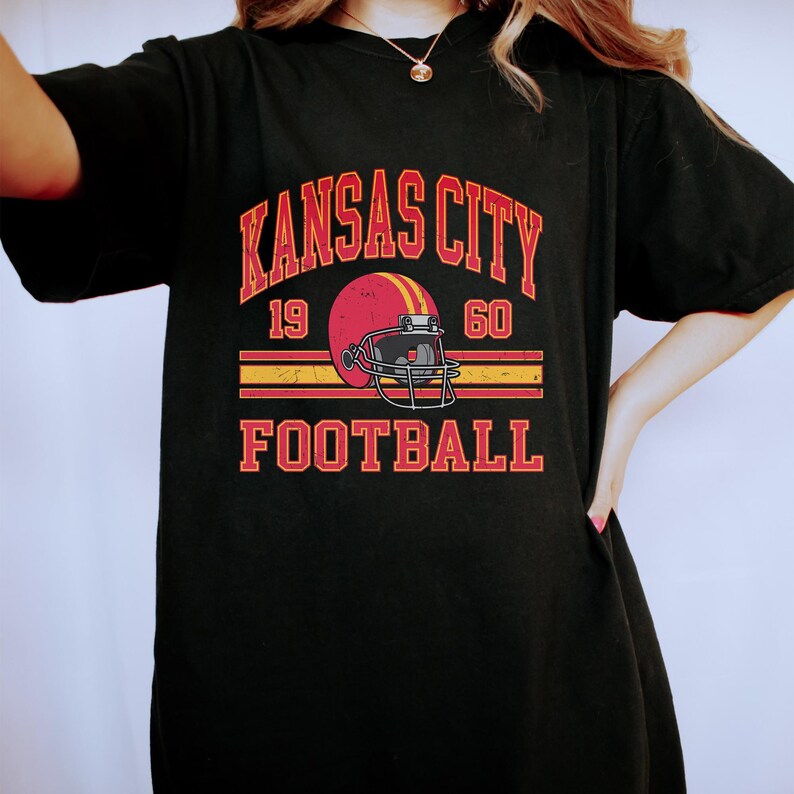 K&ouml;nnte beinhalten: Schwarzes T-Shirt mit einem Grafikdruck im Vintage-Stil eines Footballhelms und dem Text "Kansas City Football" mit den Zahlen 19 und 60.