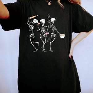 Op de afbeelding: Zwart oversized T-shirt met een afbeelding van drie skeletten die verkleed zijn als koks. De skeletten houden een deegroller, een garde, een cupcake en een kom vast.