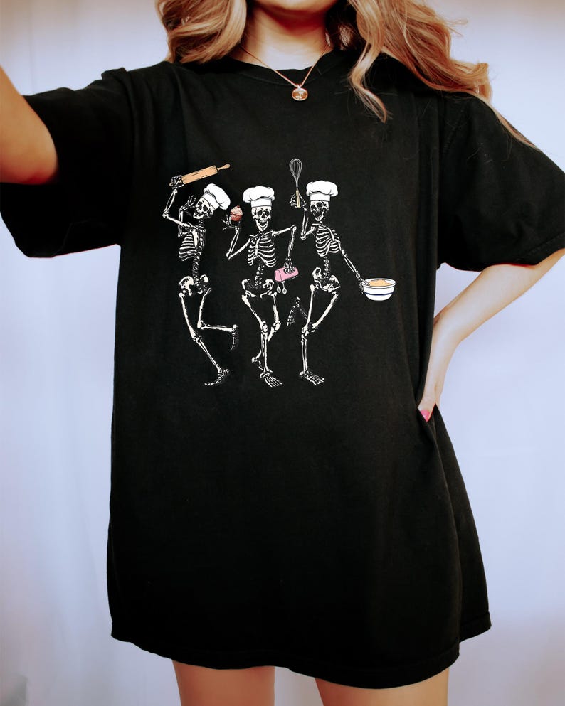 Op de afbeelding: Zwart oversized T-shirt met een afbeelding van drie skeletten die verkleed zijn als koks. De skeletten houden een deegroller, een garde, een cupcake en een kom vast.