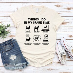 May include: A beige t-shirt with a black graphic that says "Things I do in my spare time" and shows a series of illustrations of a chihuahua. The illustrations show a chihuahua being petted, a chihuahua being looked at, a chihuahua being researched on a laptop, a chihuahua being talked about, a chihuahua being thought about, and a chihuahua being dreamed about.