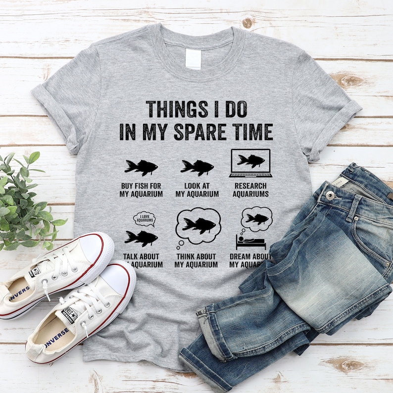 May include: A gray t-shirt with the text "Things I do in my spare time" and a series of illustrations of fish and aquariums. The illustrations depict buying fish, looking at an aquarium, researching aquariums, talking about an aquarium, thinking about an aquarium, and dreaming about an aquarium.