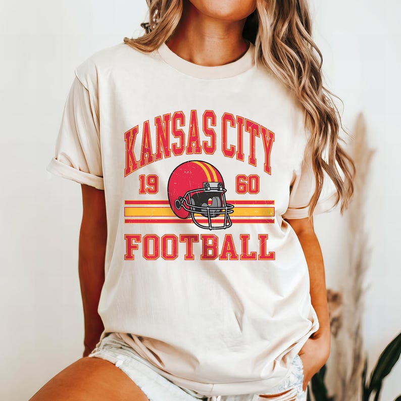 K&ouml;nnte beinhalten: Ein cremefarbenes T-Shirt mit einer Grafik im Vintage-Stil eines Footballhelms und dem Text "Kansas City 19 60 Football".