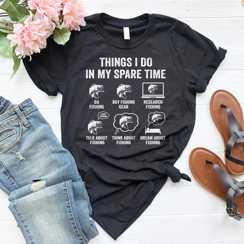 May include: A black t-shirt with a white graphic that says "Things I do in my spare time" and lists activities related to fishing. The graphic includes illustrations of fish, fishing gear, and a laptop.