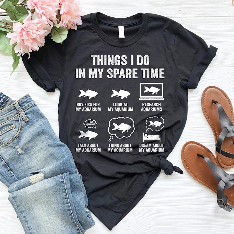 May include: Black t-shirt with white text that reads "Things I do in my spare time". The text is surrounded by illustrations of fish and aquariums. The illustrations depict activities like buying fish, looking at aquariums, researching aquariums, talking about aquariums, thinking about aquariums, and dreaming about aquariums.