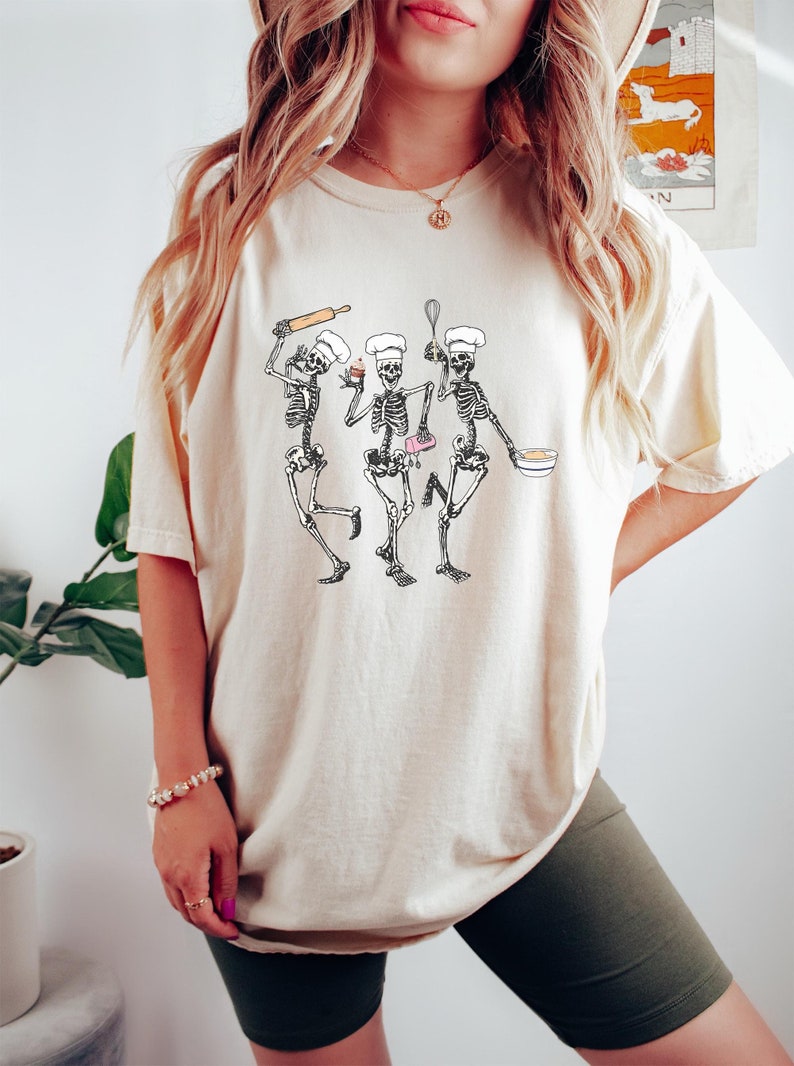 Op de afbeelding: Een wit T-shirt met een afbeelding van drie skeletten die verkleed zijn als koks en aan het bakken zijn. De skeletten houden een deegroller, een cupcake, een mengkom en een garde vast.
